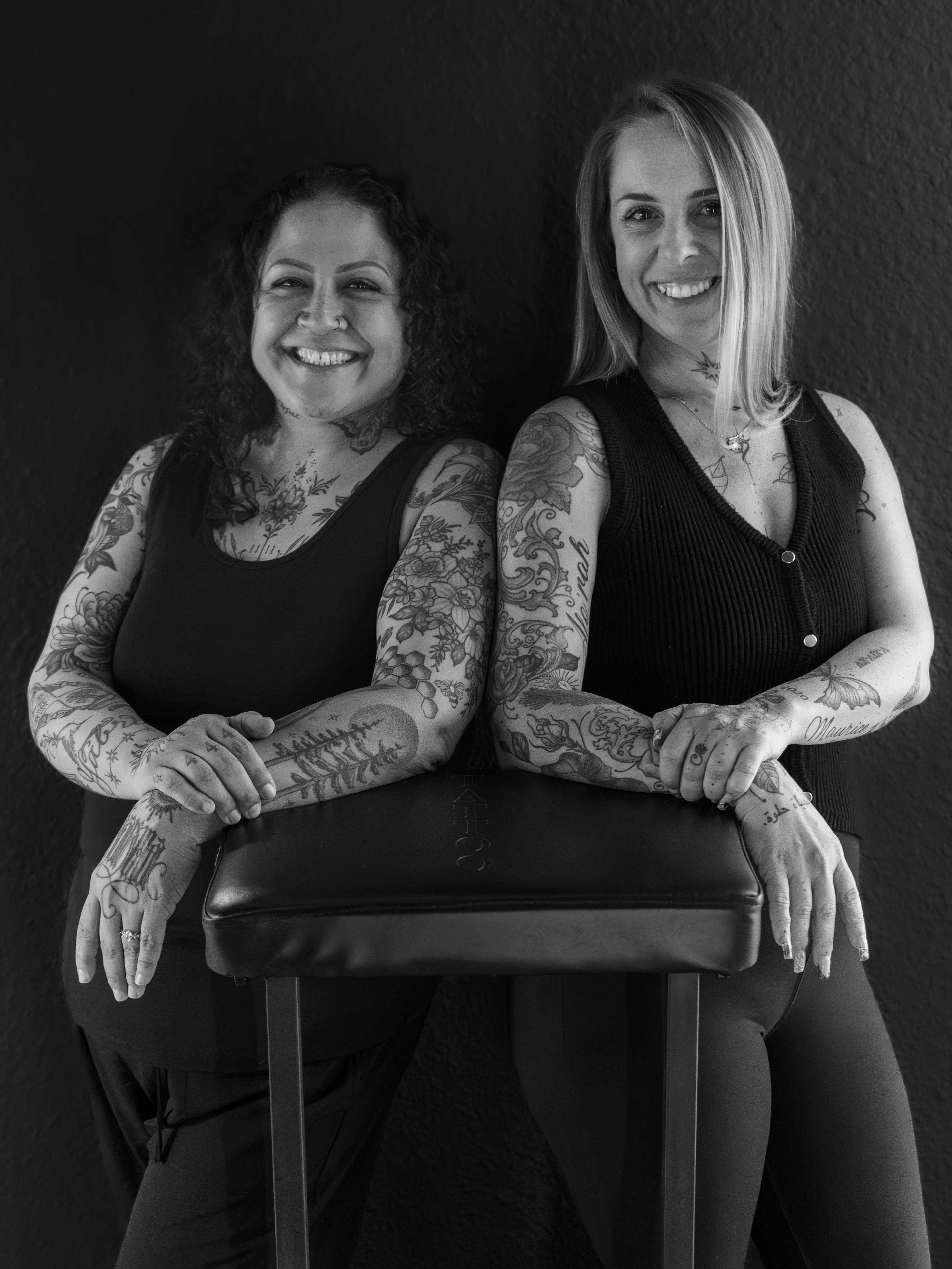 Two tattooed artists leaning on a tattoo armrest, smiling